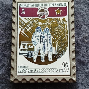 May include: Vintage postage stamp featuring two astronauts in space suits, a vehicle, and industrial structures. The stamp has text in Russian, the year 1980, and a value of 6. The stamp is rectangular with a gray border and a maroon and gold color scheme.