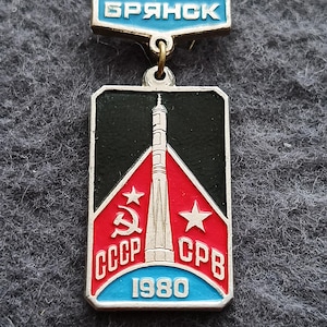 May include: A vintage pin with a rectangular design featuring a rocket, stars, and the text "CCCP" and "1980". The pin has a blue and red color scheme with the word "Брянск" at the top.