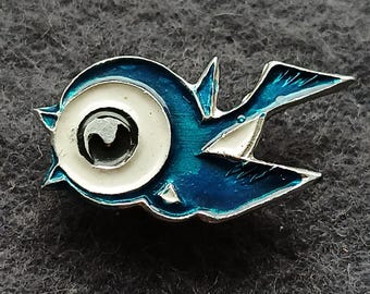 May include: A blue and white enamel bird pin with a silver-colored outline. The bird has a large eye with a black pupil and a white iris. The wings and body are blue with white accents. The pin is set against a gray background.
