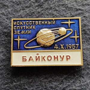 Puede incluir: Pin rectangular con fondo azul y detalles dorados. Presenta una representación dorada de un planeta con un anillo, el texto "Искусственный Спутник Земли" y "4.X.1957". La parte inferior del pin dice "Байконур".