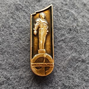 May include: A vintage gold-toned pin featuring a spaceman figure. The pin has a rectangular shape with a rounded top. The spaceman stands on a circular emblem with Cyrillic text. The pin is likely a collectible item.
