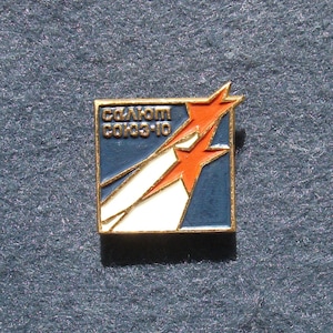 May include: A square pin with a gold border featuring a blue background with white and orange starbursts and text. The text reads "Союз Салют Союз-10" in gold lettering. The pin is likely a vintage collectible.