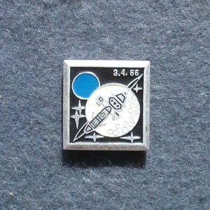 Puede incluir: Pin cuadrado plateado con un diseño espacial. El pin tiene un fondo negro con un círculo azul y una luna blanca con una nave espacial. La fecha "3.4.66" está impresa en la parte superior. También hay cuatro estrellas plateadas.