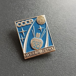Puede incluir: Un pin de esmalte azul y dorado con el texto "CCCP" y "ЛУНА 20 ЛУННЫЙ ГРУНТ" en escritura cirílica. El pin representa un cohete que se lanza al espacio con una luna y estrellas en el fondo.