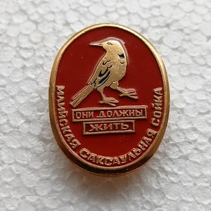 Può includere: Spilla ovale con un'illustrazione di uccello dorato su sfondo rosso. La spilla ha bordi dorati e testo in lingua straniera. L'uccello è raffigurato con le ali leggermente aperte. Il testo recita "Devono vivere."