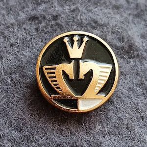 May include: Round, gold-rimmed pin with a black center featuring a gold crown and stylized wings. The bottom section is split between white and black. The pin is set against a gray, textured background.
