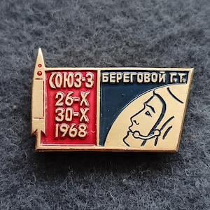 May include: Vintage Soviet-era pin with a gold border. The pin features a red and blue section with Cyrillic text and the dates 26-X and 30-X 1968. A profile of a cosmonaut is on the right side.