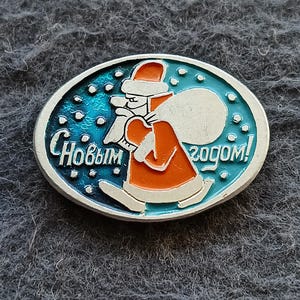 Puede incluir: Un broche ovalado de metal con una figura con abrigo y gorro rojos que lleva un saco blanco sobre un fondo azul con puntos blancos que representan nieve. El texto cirílico "С Новым годом!" está grabado en color plateado.