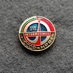 May include: A round, vintage Interkosmos pin with a red, blue, and gold design. The pin features the text "INTERKOSMOS" and "CCCP-НРБ" with the date "10.04 1979". A hammer and sickle are visible at the top.