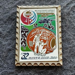 May include: A rectangular commemorative pin featuring a 1980 Soviet postage stamp design. It displays two astronauts, a space capsule, a helicopter, and the Interkosmos logo. Text includes "МЕЖДУНАРОДНЫЕ ПОЛЕТЫ В КОСМОС", "ИНТЕРКОСМОС СССР-CUBA", and "32 ПОЧТА СССР 1980".