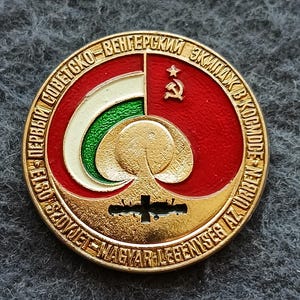 May include: A circular gold-toned metal commemorative pin featuring a red background with a hammer and sickle symbol. It displays a stylized white and green curved graphic, a central gold orb, and a black silhouette of a space station. Text in Russian and Hungarian encircles the design.