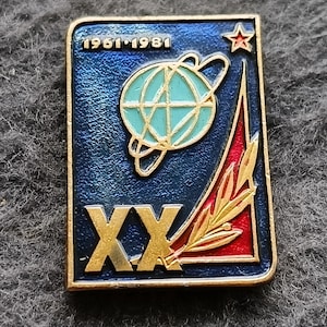 Puede incluir: Pin vintage con fondo azul y borde dorado. El pin presenta el texto "1961-1981" y "XX" en dorado. También hay un globo terráqueo, una estrella y un diseño rojo y dorado. El pin es rectangular.