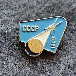 Puede incluir: Pin de esmalte vintage con fondo azul claro, las letras "CCCP" y el año "1959". El pin representa una nave espacial y un planeta dorado, con un borde dorado.