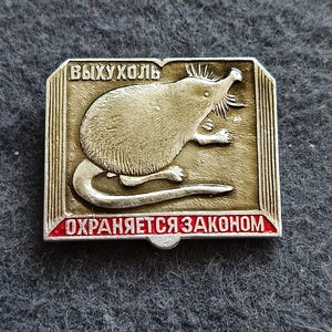 May include: A vintage silver-toned rectangular pin depicting a stylized mole-like animal. The pin features Cyrillic text: "ВЫХУХОЛЬ" at the top and "ОХРАНЯЕТСЯ ЗАКОНОМ" at the bottom, with a red border.