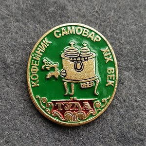 May include: Oval-shaped green and gold pin featuring a samovar design. The pin has text in a circular pattern around the edge, with the words "SAMOBAR" and "XIX BEK" visible. The central image is a gold samovar.
