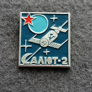 Puede incluir: Pin cuadrado vintage con temática espacial. El pin presenta un fondo azul con un satélite, una estrella roja y el texto "CAЛЮT-2" en plata. El pin tiene un borde plateado.
