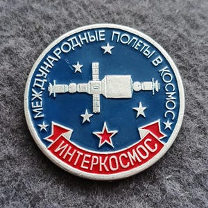 May include: A round, blue and red pin with a silver border. The pin features a stylized space station design with the text "МЕЖДУНАРОДНЫЕ ПОЛЕТЫ В КОСМОС" and "ИНТЕРКОСМОС" in white lettering. Several white stars are also present.