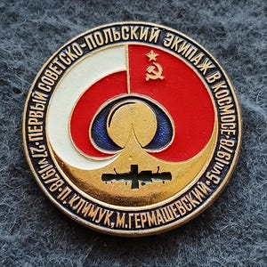 May include: A circular metal commemorative pin featuring a red and white enamel design with a gold star and hammer and sickle emblem. The center displays a stylized sun and space station graphic. Cyrillic text encircles the border of the badge.