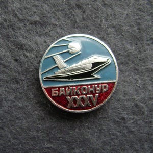 Puede incluir: Pin redondo con borde plateado. Presenta un diseño de transbordador espacial sobre un fondo azul y rojo. El texto "БАЙКОНУР XXXV" en cirílico se muestra en la parte inferior roja.