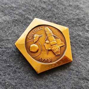 Puede incluir: Un pin vintage dorado con forma pentagonal. El pin presenta un emblema circular con un diseño de cohete y satélite, junto con texto cirílico. Las palabras "Sputnik" y "Luny" son visibles. El pin tiene un acabado metálico brillante.