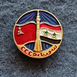 May include: A circular gold-tone metal pin featuring a central rocket design flanked by a red flag with a gold hammer and sickle and a Syrian flag with two green stars. The text CCCP and Arabic script are inscribed in red along the bottom edge.