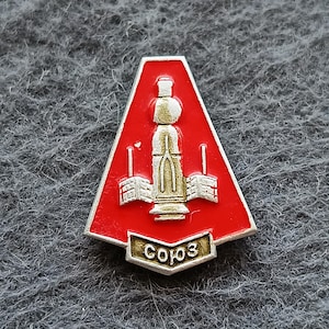 Puede incluir: Un pin de metal trapezoidal con un fondo de esmalte rojo vibrante. En el centro, una representación en relieve de color plateado de una nave espacial con paneles solares extendidos. En la base, una pequeña banda muestra el texto cirílico "Союз".