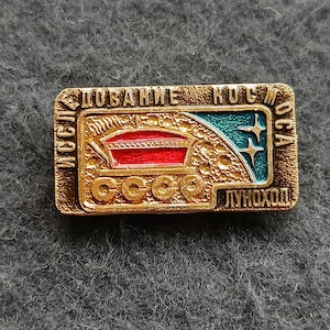 Puede incluir: Pin rectangular vintage con marco de metal dorado. El pin presenta un diseño rojo y verde azulado con el texto "CCCP" y texto cirílico. El pin representa un diseño temático espacial con estrellas y un vehículo.