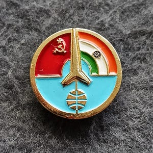 May include: A round, gold-rimmed pin with a design featuring red, orange, blue, and green sections. The pin includes a hammer and sickle, a stylized arrow, and a wheel symbol, with a gold-colored border.