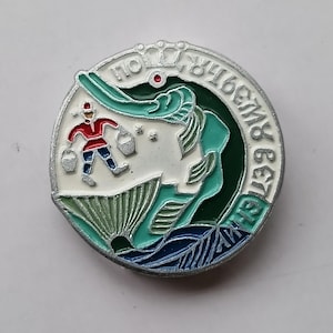 Puede incluir: Un pin redondo esmaltado con borde plateado. El diseño presenta una figura con ropa roja que lleva cubos, una cara estilizada y texto en un patrón circular. Los colores incluyen blanco, verde, azul y rojo.