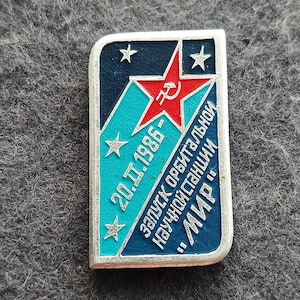 以下が含まれることがあります： 銀色の縁取りのある長方形のピンバッジ。ハンマーと鎌の付いた赤い星、青とターコイズの背景、白で「20.11.1986」と「MIR」の文字が特徴です。ピンバッジには白い星も3つあります。