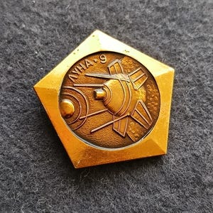 Puede incluir: Un pin dorado vintage con forma pentagonal. El pin presenta un emblema circular con un diseño de nave espacial y el texto cirílico "ЛУНА-9". El pin tiene un diseño detallado en relieve.