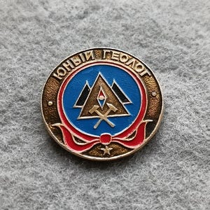 May include: A round, vintage pin with the Cyrillic text "ЮНЫЙ ГЕОЛОГ" in gold lettering. The pin features a blue center with a mountain and pickaxe design, surrounded by a red ribbon and a gold border.
