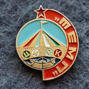 May include: Vintage enamel pin with a circus tent design in gold, teal, red, and white. The pin features a red star and a semi-circular red border with gold lettering. A collectible item.