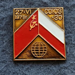 May include: A square metal pin featuring a bronze background with a central chevron design in red and white enamel. The pin displays a gold hammer and sickle symbol, a globe icon, and the text 27 VI 1978 and COЮ3 30 in raised gold lettering.