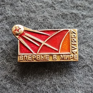 Puede incluir: Pin vintage con un diseño rojo y naranja que presenta una estrella y una trayectoria estilizada. El pin tiene detalles dorados y el texto "Впервые в мире" y "1957".