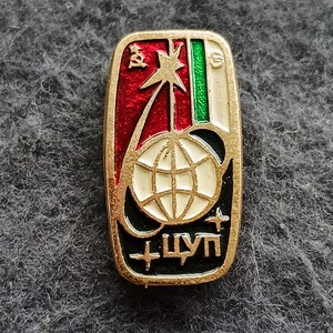 May include: A rectangular gold-toned pin with a red, white, green, and black design. The pin features a globe, a star, and Cyrillic text. The pin has a gold border.