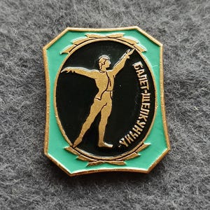 May include: A vintage pin featuring a gold-colored ballet dancer on a black background, surrounded by a green border with gold trim. The pin has Cyrillic text. The pin is rectangular with rounded corners.