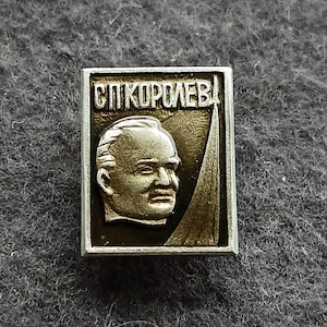 May include: A rectangular silver-toned pin featuring a portrait of a man and the Cyrillic text "СП.КОРОЛЕВ". The portrait is set against a dark background with a stylized element. The pin has a silver border.