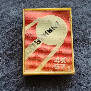 Puede incluir: Un broche rectangular con borde color crema sobre un fondo rojo con estrías horizontales. Un gráfico estilizado de color crema con forma curva que recuerda a una hoja o una hoz está centrado, con caracteres japoneses en la esquina inferior derecha.