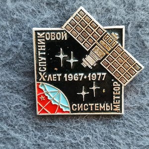 Puede incluir: Un pin metálico cuadrado con fondo negro, un satélite plateado, estrellas de cuatro puntas y un segmento de globo rojo y azul claro. El texto "Спутник", "1967-1977", "Метеор" y "Системы" aparece en letras plateadas sobre el esmalte.