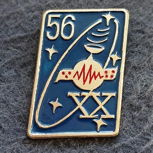 Puede incluir: Un pin rectangular de metal dorado con fondo de esmalte azul oscuro. Presenta el número 56, el número romano XXV y un icono de satélite dorado central con una línea roja en zigzag. Estrellas de cuatro puntas rodean el diseño central.
