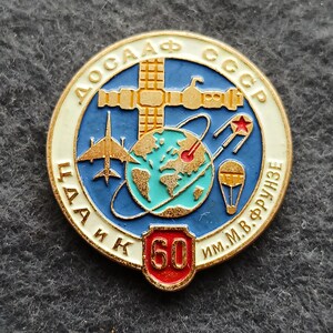 May include: A round, vintage pin with a blue and white border. The pin features a gold structure, a globe, a jet, a satellite, and a parachute. The center of the pin has the number 60 in red.