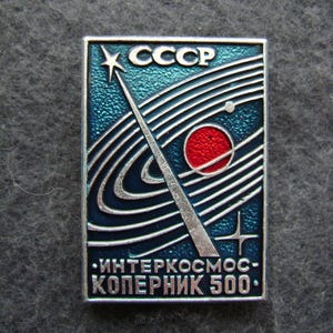 May include: A rectangular pin with a blue background and silver border. The pin features the text "CCCP" at the top, with a star and a stylized depiction of a planet and orbits. The text "ИНТЕРКОСМОС-КОПЕРНИК 500" is at the bottom.