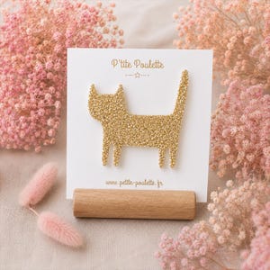 Peut inclure: Une broche en forme de chat à paillettes dorées fixée sur une carte de présentation carrée blanche. La carte porte le texte P'tite Poulette en haut et www.petite-poulette.fr en bas. La carte est maintenue par un support en bois cylindrique.