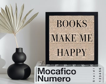 May include: A black framed print with the words "BOOKS MAKE ME HAPPY" in bold black letters. The background of the print is a collage of text. The print is displayed on a white surface, next to a black vase and a white book.
