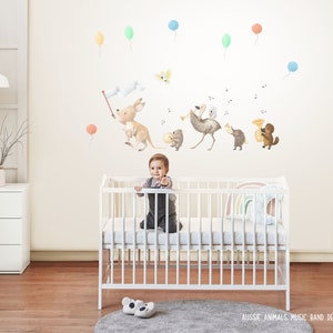 Peut inclure: Un lit bébé blanc avec un bébé à l'intérieur, entouré de ballons colorés et d'autocollants muraux représentant des animaux australiens jouant de la musique. Les autocollants représentent un kangourou, un émeu, un wombat et un ornithorynque. Le texte "Aussie Animals Music Band Decals - Facing Left" est visible en bas de l'image.