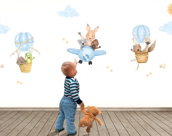 May include: A child points at a blue airplane with a kangaroo, koala, and echidna on board. Two hot air balloons with animals are on the wall behind the child. The balloons are blue and white with yellow and orange accents. The wall is white.