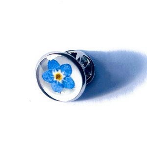 May include: A round, silver-toned lapel pin featuring a blue forget-me-not flower encased in clear resin. The flower has a yellow center and is set against a white background. The pin has a classic, simple design.