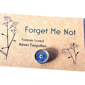 May include: A Forget Me Not pin with a real blue flower encased in clear resin, set on a silver-colored metal backing. The pin is displayed on a tan card with the words "Forget Me Not" and "Forever Loved, Never Forgotten".