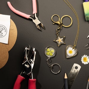 May include: An overhead shot of craft supplies and handmade jewelry. Items include a star-shaped keychain, a heart-shaped daisy pendant, a four-leaf clover keychain, and tools like pliers and a hole punch. The image also shows the text "Cotswold Crafts by Emma" on a sticker.